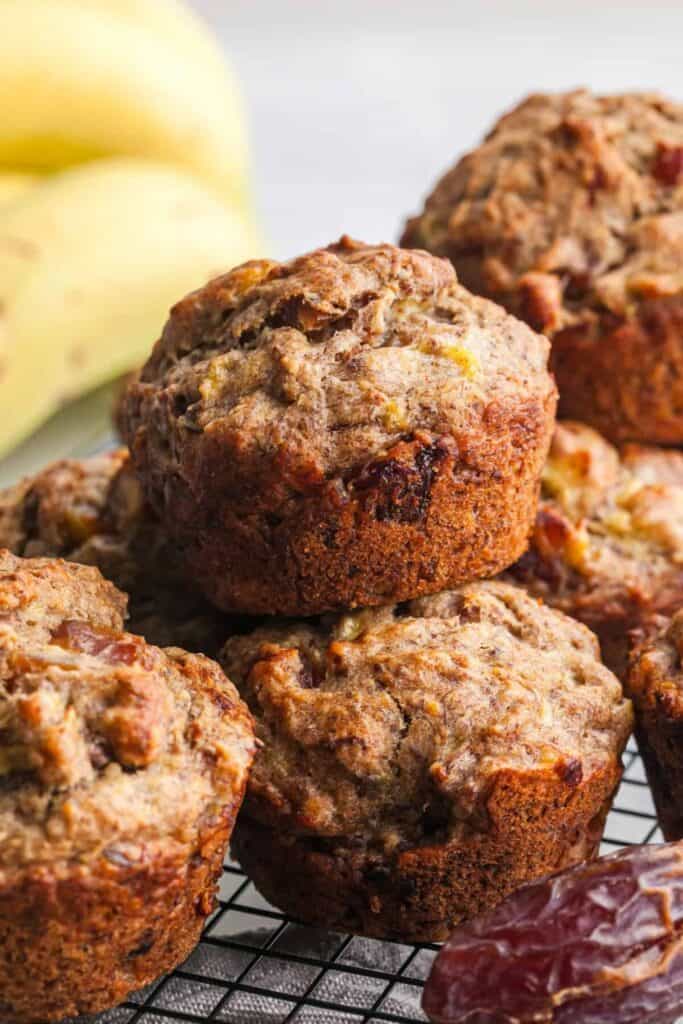Close up of banana date muffins cooling on a wire rack, with visible chunks of chopped dates baked into the moist crumb. The tops are golden brown and slightly domed, showing a hearty texture from ripe bananas and whole ingredients.