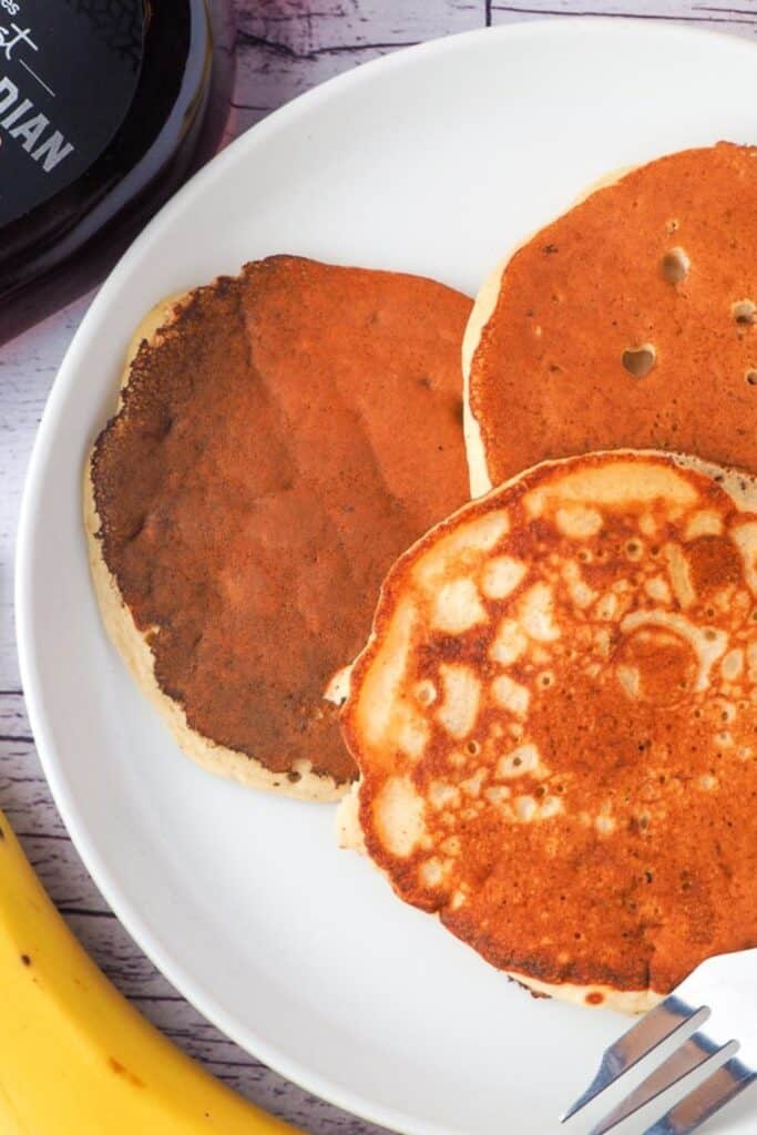 Plate of banana pikelets with golden brown tops and slightly crisp edges, showing one darker pancake alongside lighter fluffy ones. The pikelets sit on a white plate with a fork nearby and a whole banana in the background.