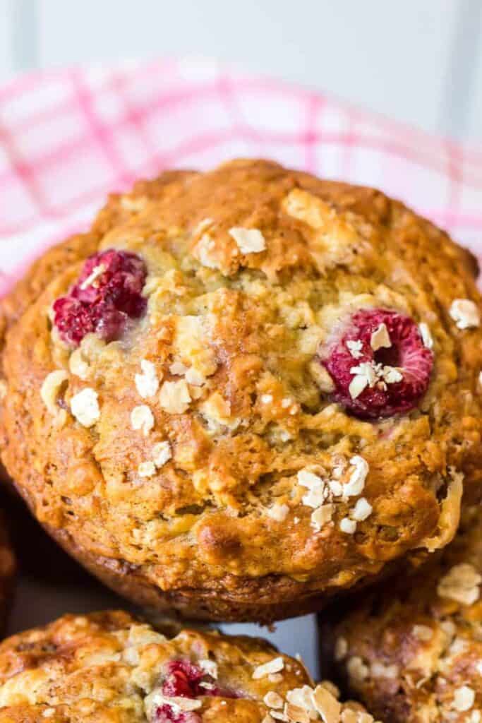 Close-up of a golden banana raspberry muffin with white chocolate chips, topped with oats and studded with whole raspberries. The muffin has a moist, hearty texture and sits against a pink checkered cloth backdrop for a cozy, homemade feel.