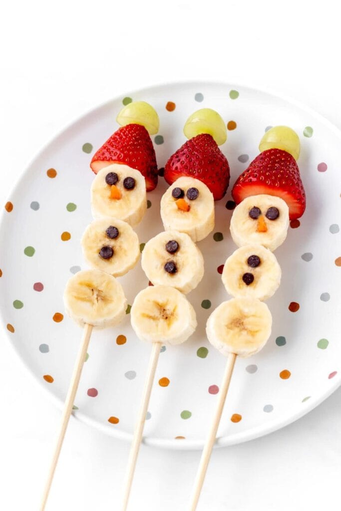 Three banana snowman on sticks, with strawberry  grape hats. 