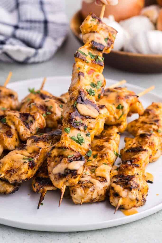 Grilled Bang Bang chicken skewers stacked on a serving plate and drizzled with creamy orange sauce. The chicken has charred edges and a sprinkle of fresh herbs.