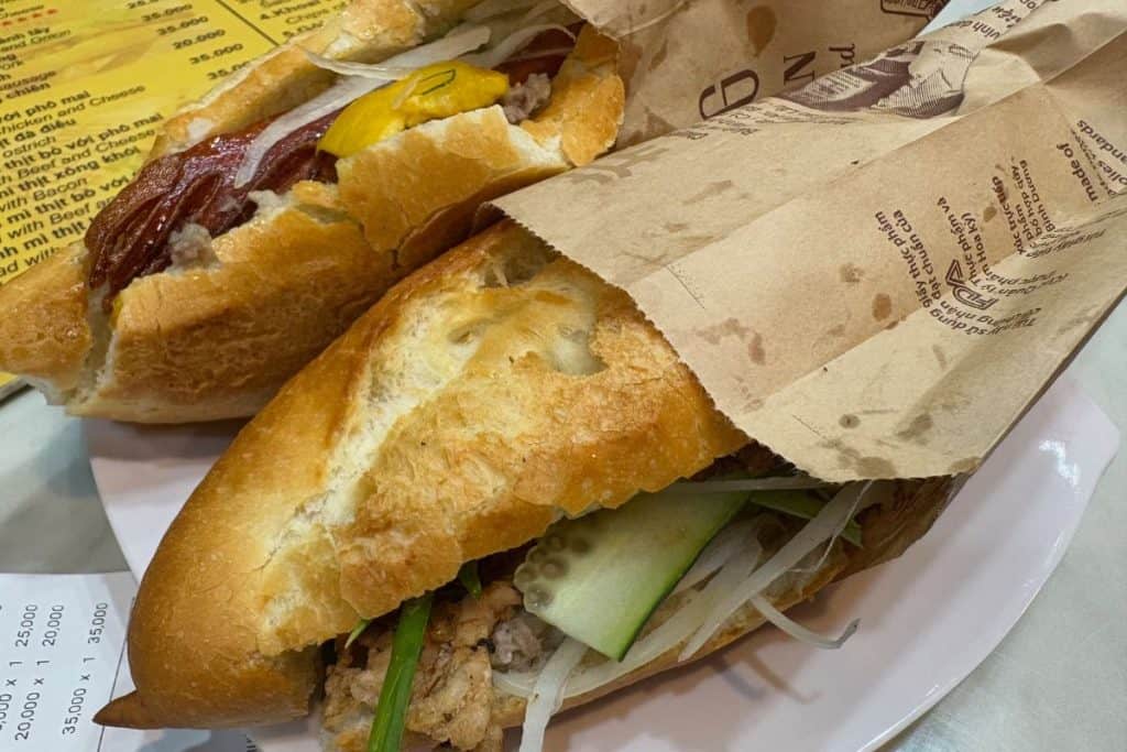 Two Banh Mi sandwiches sit on a white plate, partially wrapped in brown paper, filled with ingredients like grilled meat, pickled vegetables, cucumber slices, and chili. The crispy golden baguettes are slightly cracked open, revealing the colorful, savory fillings inside.