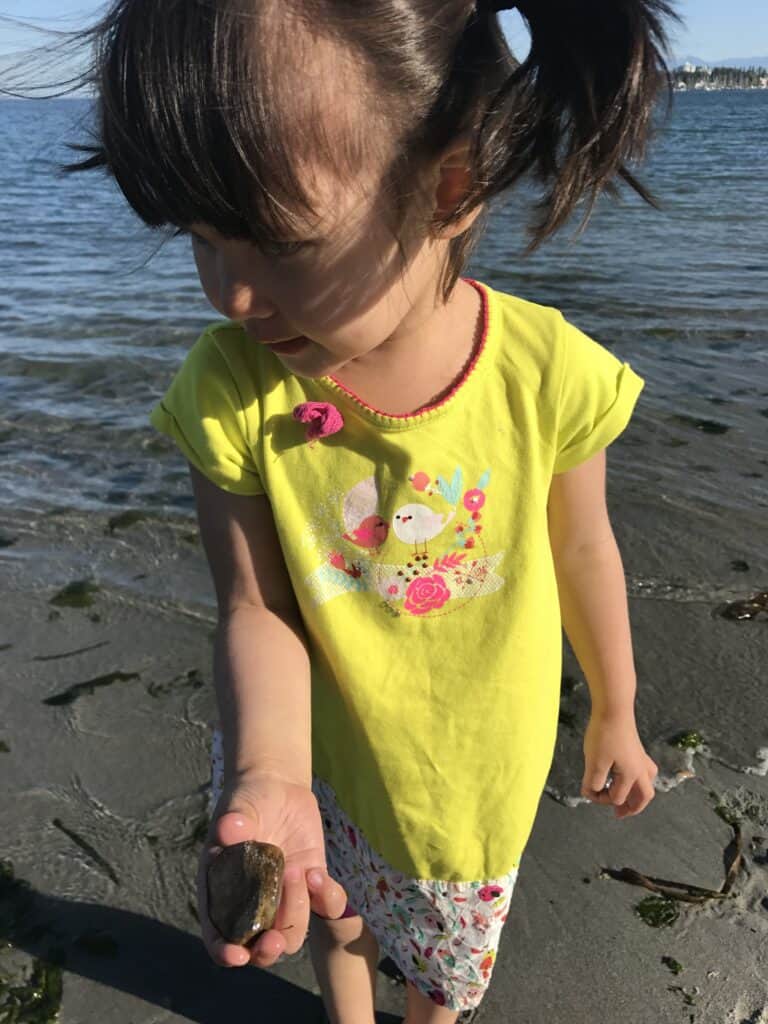 Toddler girl in brightly colored dress holding a rock at sandy beach. 