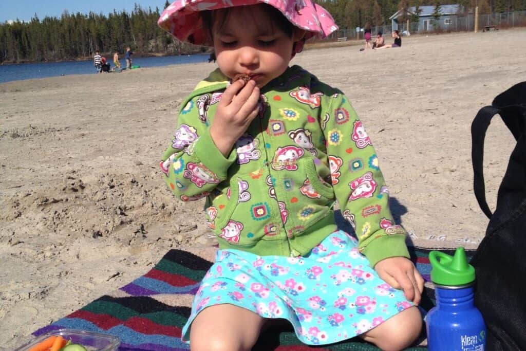 Young girl eating a snack at the beach, on a beach blanket, with reusable water bottle beside her.