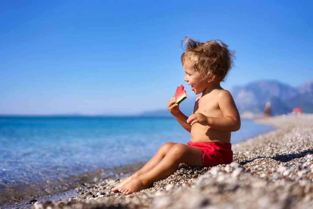 Young boy in red swimsuit eating watermelon at the beach