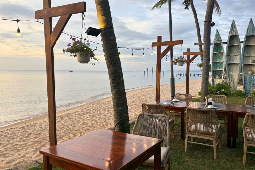 A serene beachfront dining area at Madam Keo’s in Phú Quốc, Vietnam, features wooden tables and rope-woven chairs set on grass just steps from the sandy shore. String lights and hanging flower pots add charm, while weathered surfboards stand upright near palm trees, and the calm sea stretches into the distance under a pastel sky.