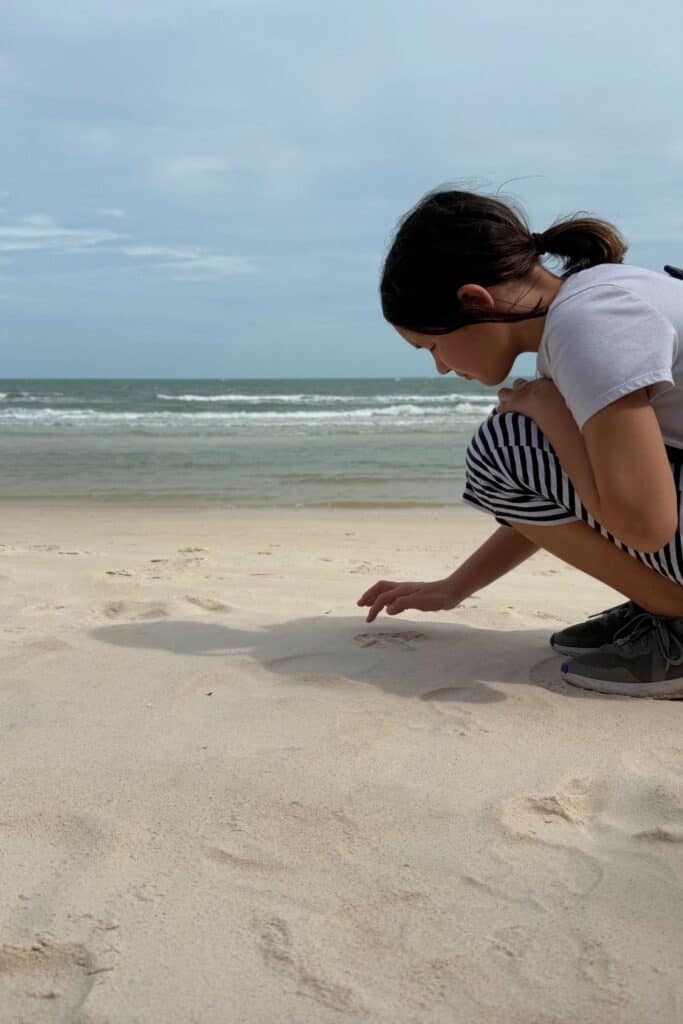 A young girl with a ponytail crouches on the soft sand of Sao Beach in Phu Quoc, carefully examining something near the shoreline with gentle waves rolling in under an overcast sky.