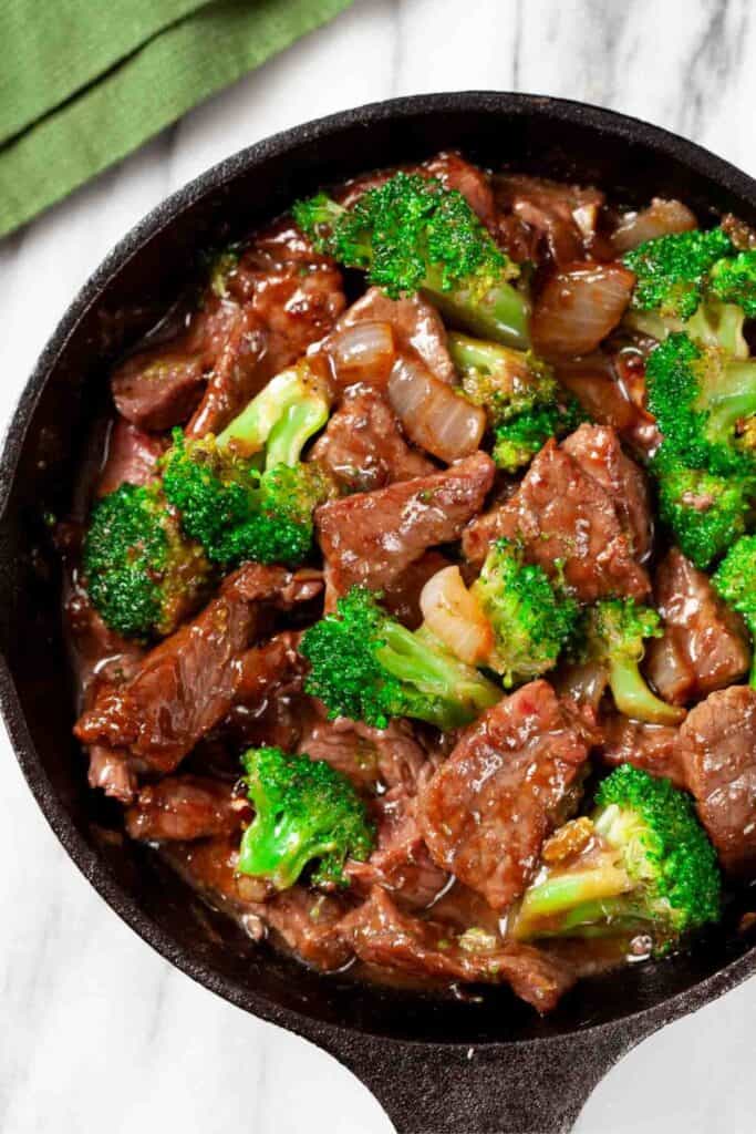 Beef and broccoli stir fry in a small cast iron skillet with tender slices of beef, bright green broccoli florets, and onions coated in a glossy brown sauce. Overhead view of this easy dinner served hot in the pan on a light marble surface with a green kitchen towel nearby.