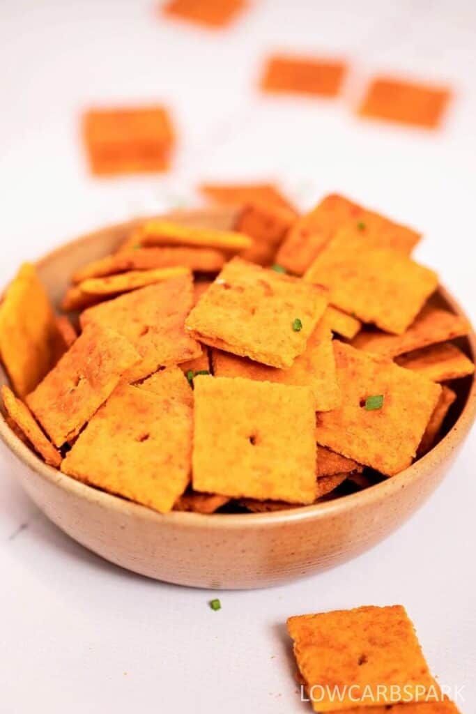 Bowl of best high protein cheddar crackers stacked high, baked to a deep golden orange color with a crisp square shape and small center hole. The crackers have a slightly rough texture with visible seasoning and a few green herb flecks on top.