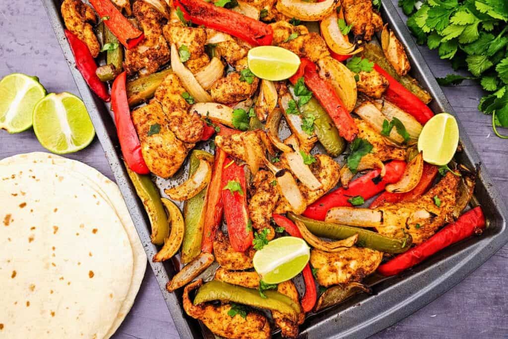 Best chicken fajitas in oven baked on a sheet pan with juicy seasoned chicken strips roasted alongside red and green bell peppers and caramelized onions. Garnished with fresh cilantro and lime wedges and served with warm tortillas for an easy flavorful dinner.