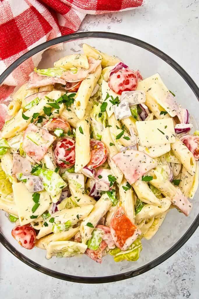 A glass bowl filled with creamy Italian pasta salad, featuring penne pasta tossed with sliced ham, pepperoni, provolone cheese, cherry tomatoes, red onion, lettuce, and chopped parsley. The dish is richly coated in a herb-speckled dressing and set beside a red and white checkered towel on a light countertop.