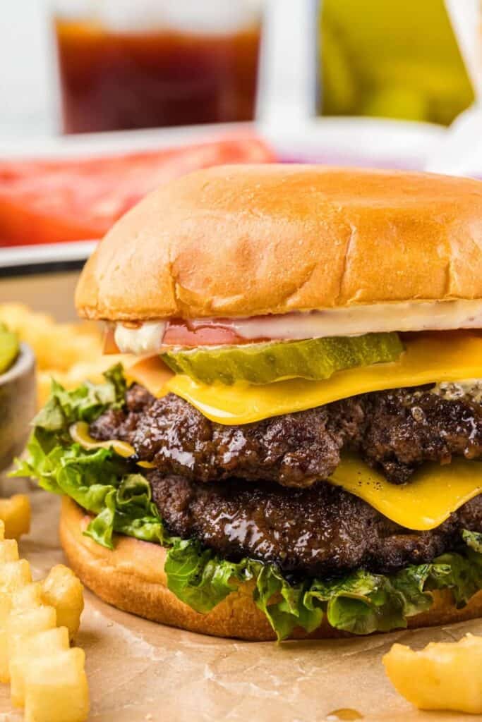 Double smashburger with two crispy beef patties, melted American cheese, pickle slices, lettuce, and sauce on a toasted bun. Served with crinkle cut fries, this hearty burger is a popular hot weather meal for backyard cookouts and casual summer dining.