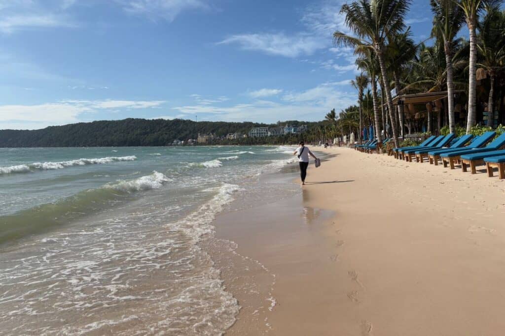 A young tween girl walking down the sandy Khem Beach in Phu Quoc, with Palm trees and beach chairs on one side.