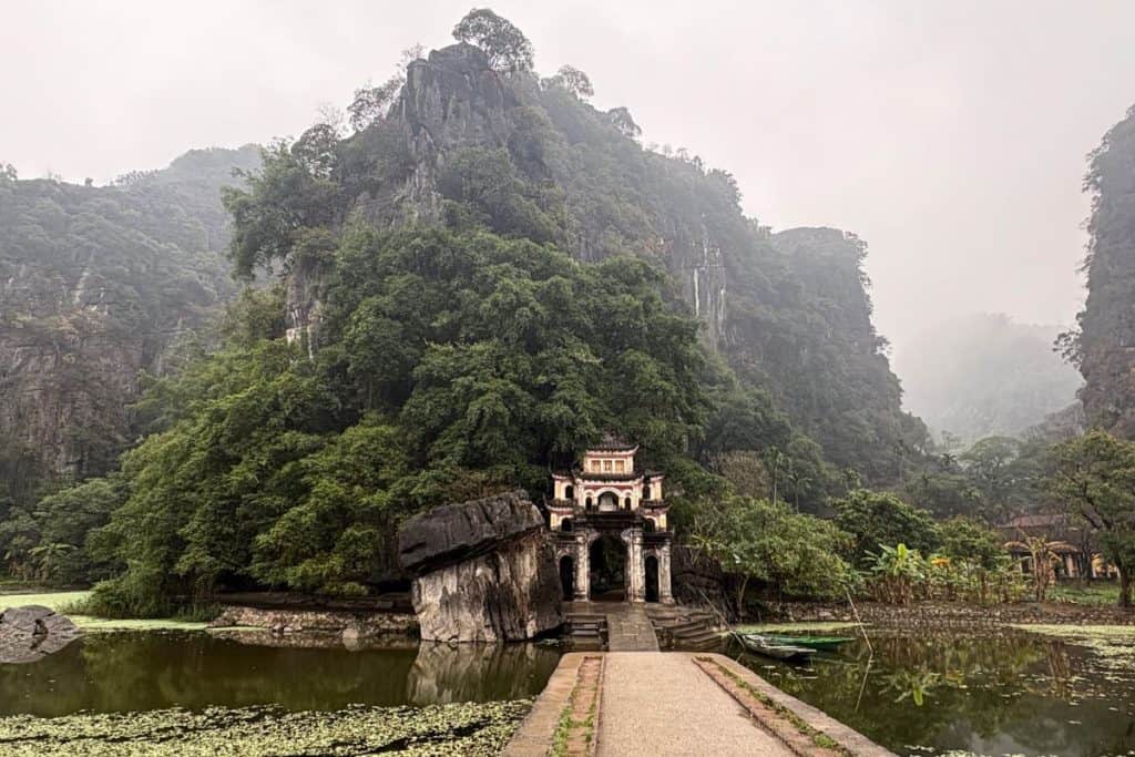 A misty mountain landscape surrounds a historic stone gate with traditional Vietnamese architecture, nestled against a lush green hillside. A narrow pathway leads across a pond filled with floating vegetation, enhancing the serene and mystical atmosphere.