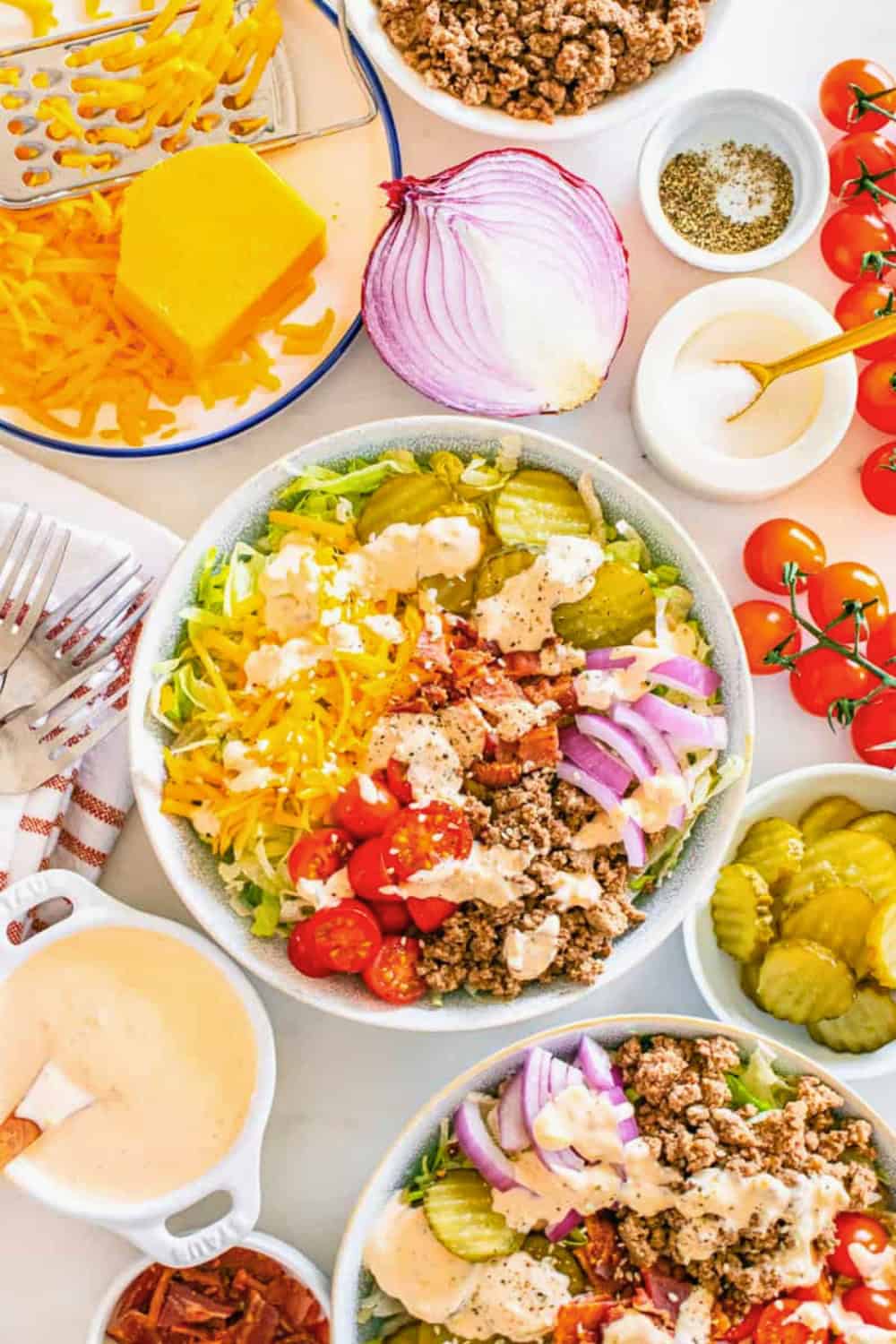 Bowl of chopped lettuce topped with seasoned ground beef, shredded cheddar cheese, bacon, cherry tomatoes, sliced pickles, red onion, crumbled cheese, and creamy burger sauce, surrounded by the fresh ingredients used to make it. These Big Mac bowls bring together all the classic burger flavors in a hearty, low-carb salad.