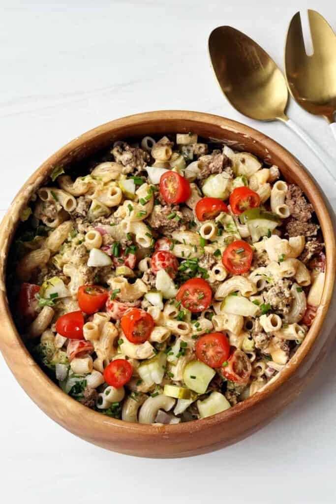 Big Mac pasta salad served in a wooden bowl, made with elbow macaroni, ground beef, chopped pickles, diced onions, cherry tomatoes, and shredded lettuce, all tossed in a creamy, tangy dressing. A pair of gold serving utensils rests beside the bowl on a white surface