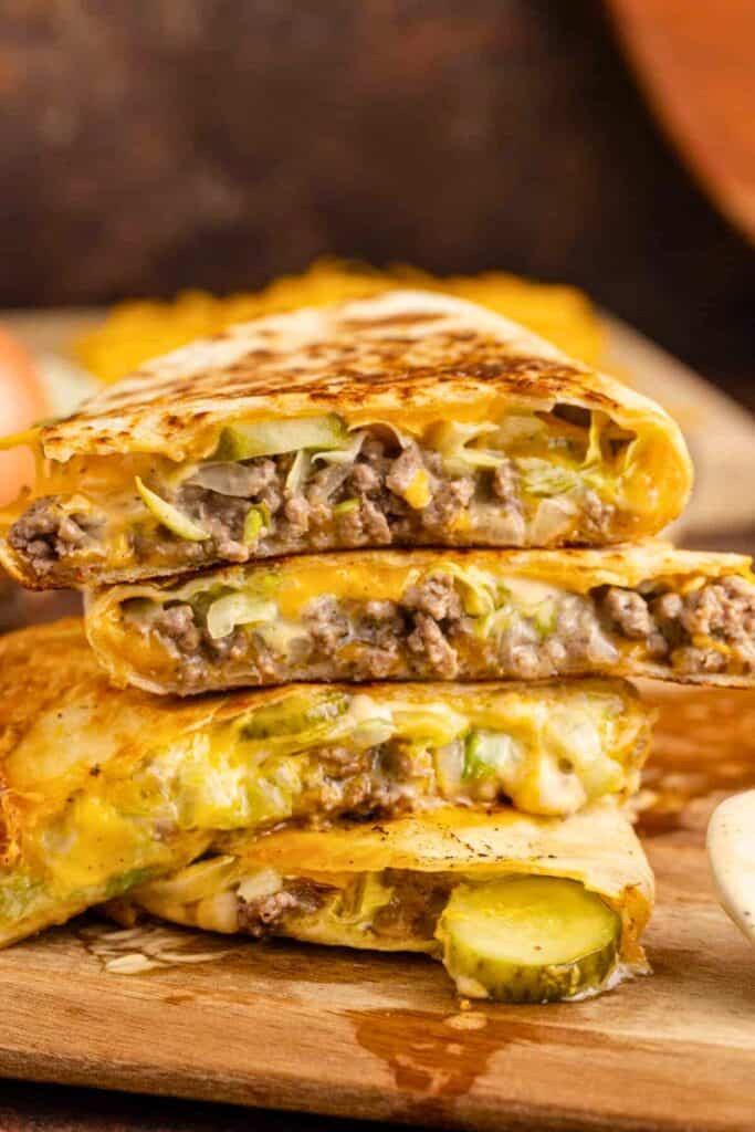 Big Mac quesadillas stacked on a wooden board with browned tortillas filled with ground beef, melted cheese, shredded lettuce, and sliced pickles. Close up of the cheesy layers and burger inspired filling spilling from the toasted quesadilla wedges.