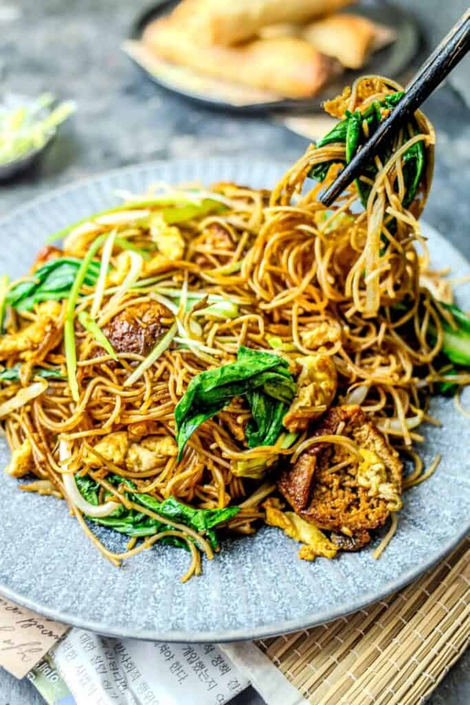 Indonesian noodles stir fried with thin noodles, egg, leafy greens, and bean sprouts tossed in a savory sauce. Chopsticks lift a bundle of noodles from the plate showing the mix of vegetables and scrambled egg.