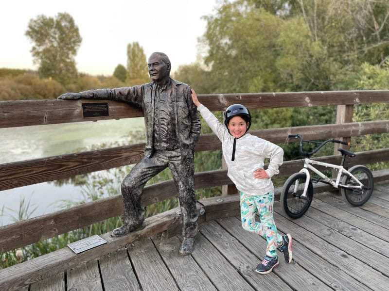 A young girl standing with her hand on a metal statue of a man, on a wooden bridge with a bike beside her. 