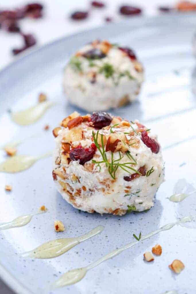 A cranberry pecan goat cheese ball topped with dried cranberries. Chopped pecans and fresh dill create a colorful easy appetizer served on a pale blue platter with a light golden honey drizzle.