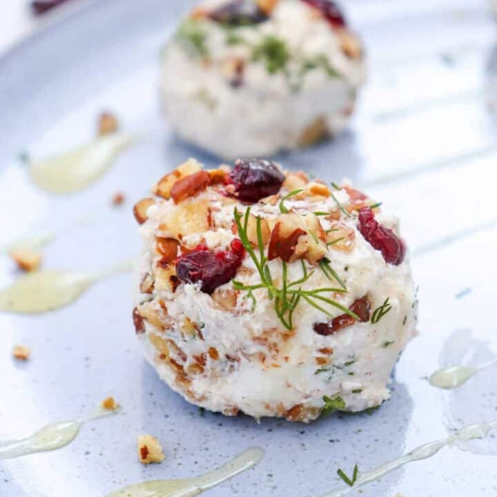 A cranberry pecan goat cheese ball topped with dried cranberries. Chopped pecans and fresh dill create a colorful easy appetizer served on a pale blue platter with a light golden honey drizzle.