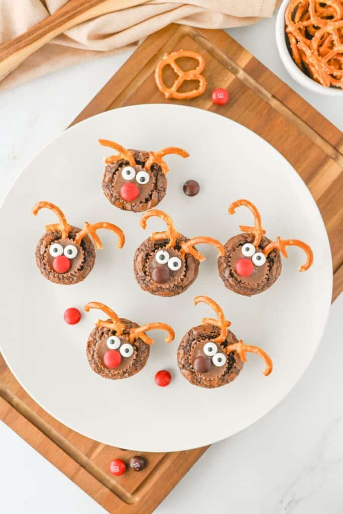Overhead view of reindeer brownies decorated with pretzel antlers, candy eyes, and red or brown candy noses arranged on a white plate. The festive Christmas treats sit atop a wooden board, surrounded by extra pretzels and scattered red and brown M&M's.
