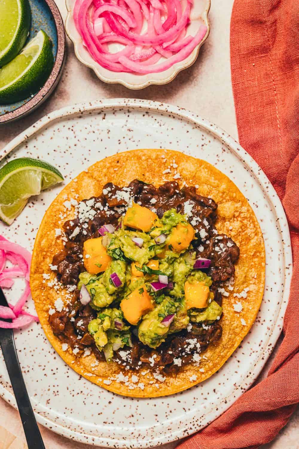Crispy corn tostada topped with seasoned black beans, chunky mango avocado salsa with diced mango and red onion, and crumbled cheese on a speckled white plate. Lime wedges and pickled red onions on the side highlight this colorful black bean tostadas recipe.