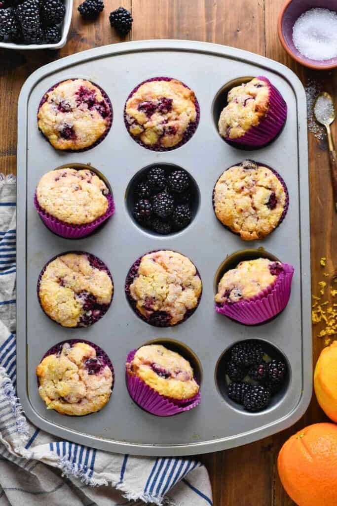 Freshly baked blackberry muffins in a muffin tin lined with bright purple wrappers, with a few cups filled with fresh blackberries. The golden tops are studded with juicy blackberries and lightly dusted with sugar, surrounded by oranges, sugar, and a rustic kitchen setup.