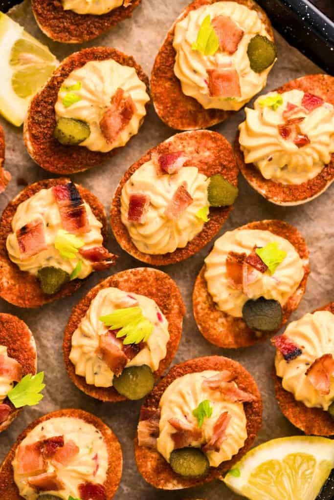 Bloody Mary deviled eggs with a deep red spiced coating, piped with creamy yolk filling and topped with bacon bits, pickle slices, and celery leaves. The deviled eggs are arranged on parchment paper with lemon wedges for garnish.