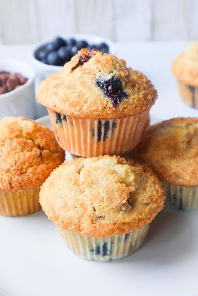 Close up of blueberry chocolate chip muffins with tall golden brown domed tops and a light sprinkle of coarse sugar. Juicy blueberries and melted chocolate chips are visible throughout the tender crumb, with small bowls of fresh blueberries and chocolate chips blurred in the background. 