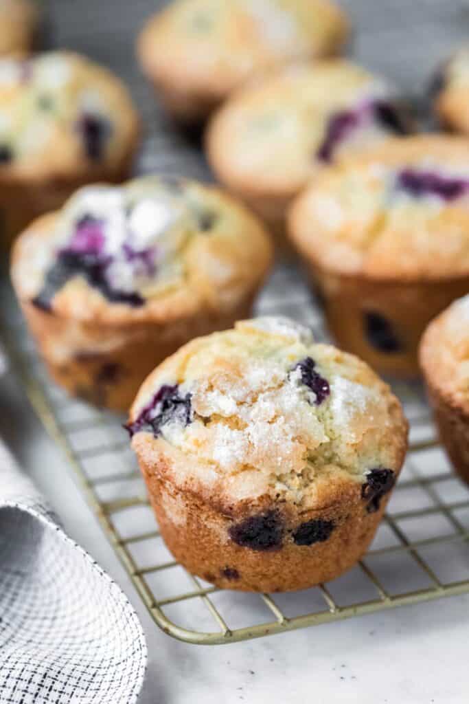 Close up of homemade blueberry lemon muffins cooling on a wire rack with golden brown tops and a coarse sugar crust. Juicy blueberries are visible throughout the fluffy crumb and bursting through the sides.
