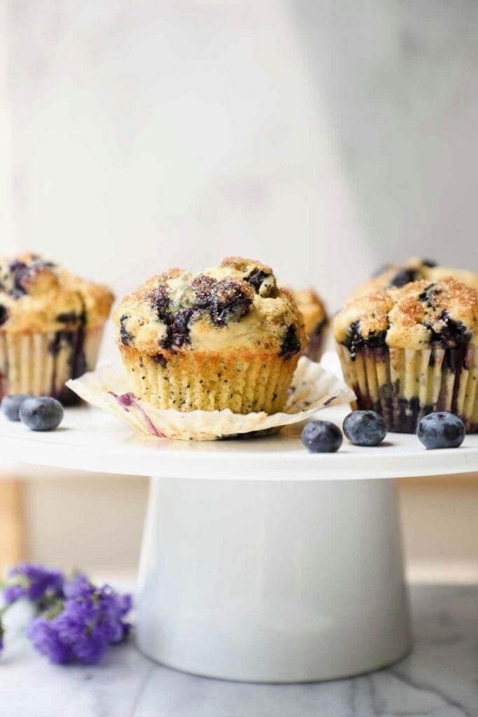 Close up of blueberry lemon poppyseed muffins displayed on a white cake stand with golden brown tops and a light sprinkle of sugar.