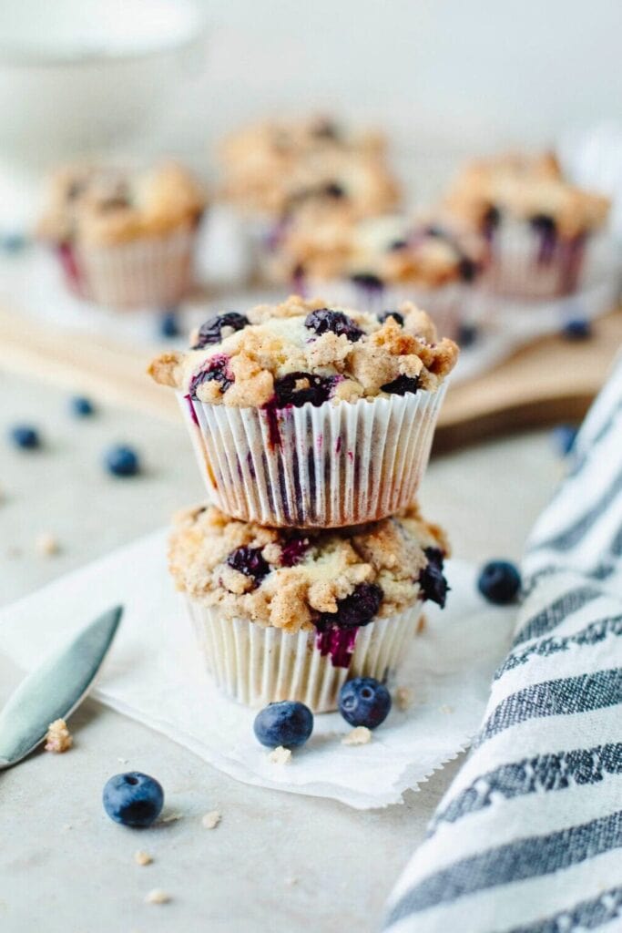 Close up of bakery style blueberry streusel muffins stacked on parchment paper with a thick crumb topping and juicy blueberries bursting through the sides. The golden brown muffins sit in white paper liners with fresh blueberries scattered around.