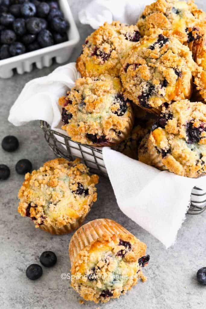 Close up of blueberry streusel muffins piled in a wire basket lined with a white cloth napkin. Each muffin has a thick crumbly streusel topping with juicy blueberries peeking through the golden brown tops, while fresh blueberries are scattered nearby.