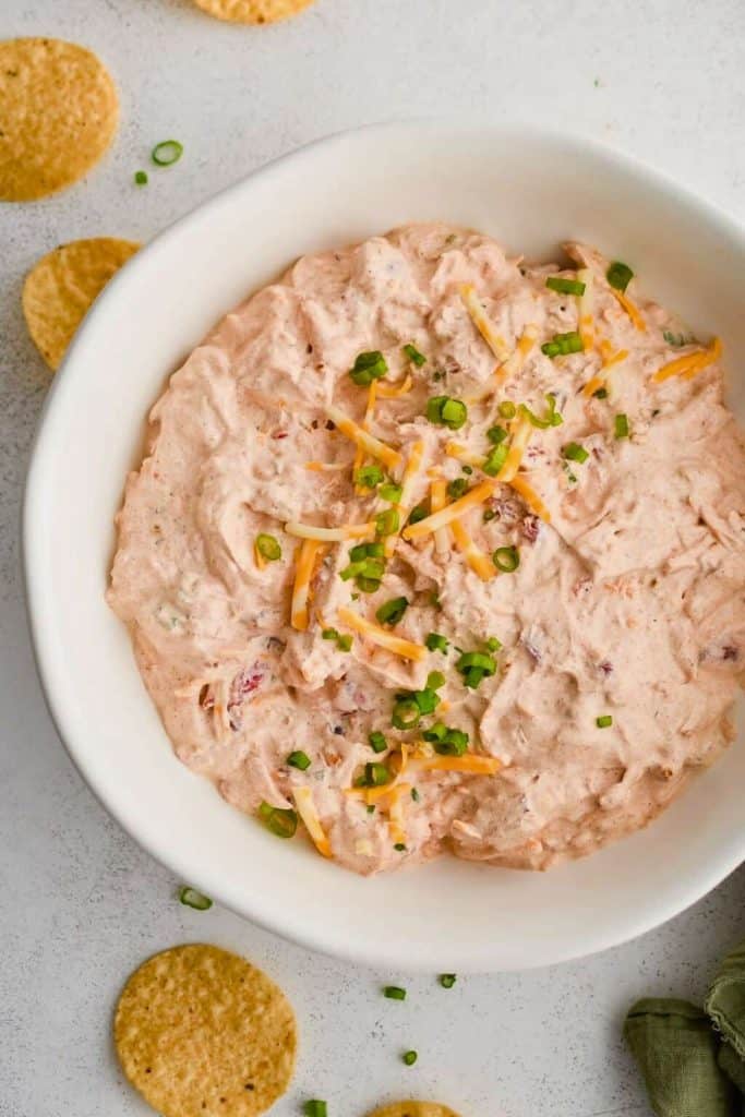 Creamy cold dip topped with shredded cheddar cheese and chopped green onions, served in a white bowl with round tortilla chips scattered around. The dip has a pinkish hue with visible bits of bacon and seasoning, perfect for tailgating.