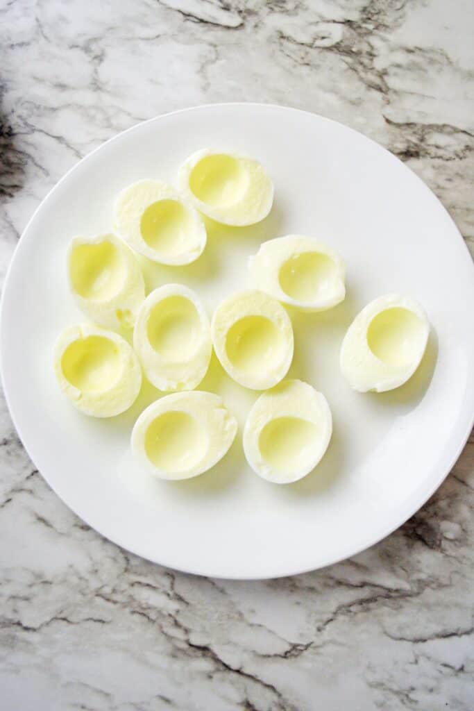 Hard boiled egg white halves with the yolks removed and arranged on a white plate. The hollow centers are ready to be filled with the seasoned yolk mixture.