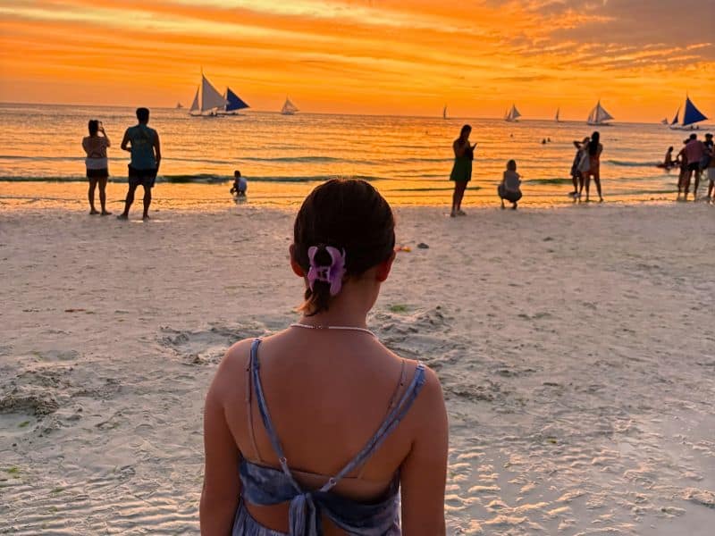 A girl with a purple hair clip stands on the white sand of Boracay beach, facing a stunning sunset over calm orange waters. Sailboats with blue and white sails glide across the horizon as beachgoers capture the view and enjoy the tranquil evening.