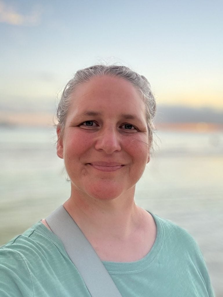 a middle aged woman with her hair pulled back is smiling at the camera with a Boracay Philippines sandy beach and sunset in the background.