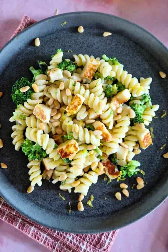 Boursin lemon pasta with rotini tossed with broccoli florets, golden seared halloumi cubes, and pine nuts on a dark plate. The pasta is lightly coated in a creamy lemon sauce with scattered pine nuts around the dish.