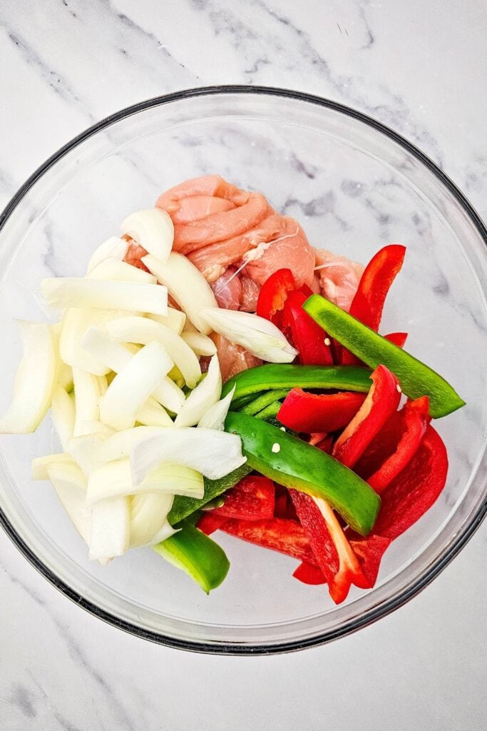 Raw chicken strips, sliced red and green bell peppers, and chopped onions combined in a clear glass mixing bowl on a marble surface. The ingredients are prepped and ready to be seasoned and baked for an easy sheet pan chicken fajitas recipe.