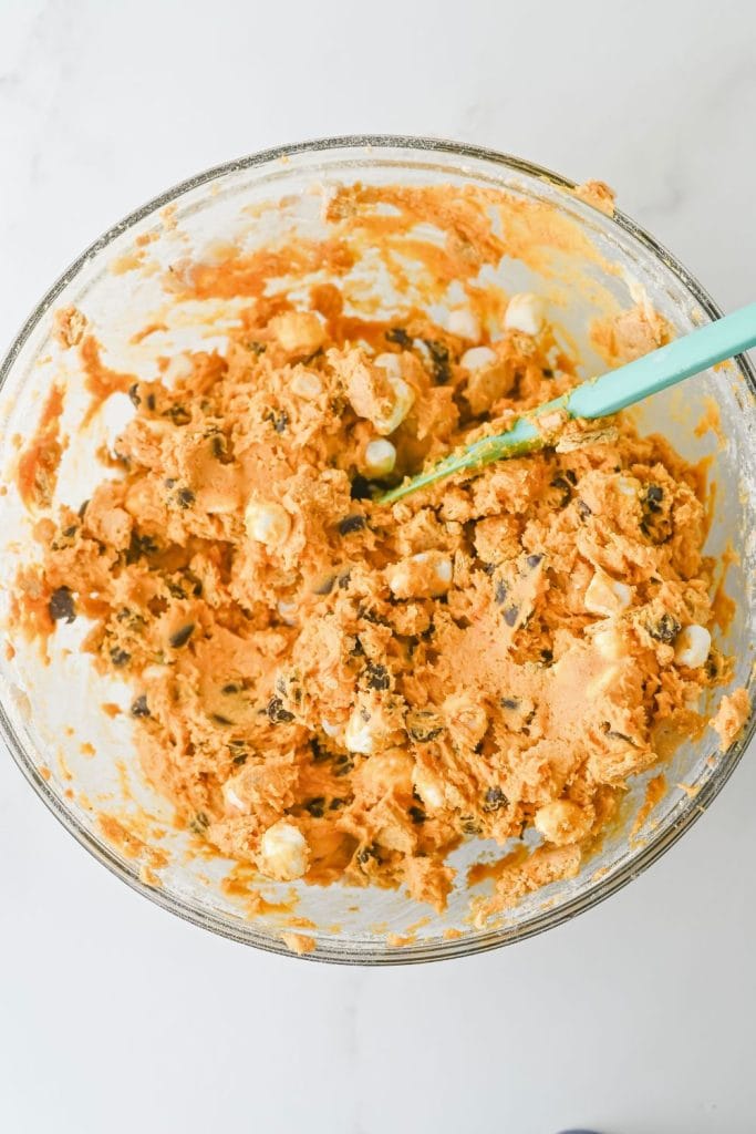  Glass mixing bowl filled with pumpkin-colored cookie dough mixed with chocolate chips and mini marshmallows. A teal spatula rests in the dough, showing it’s ready to be scooped for baking.
