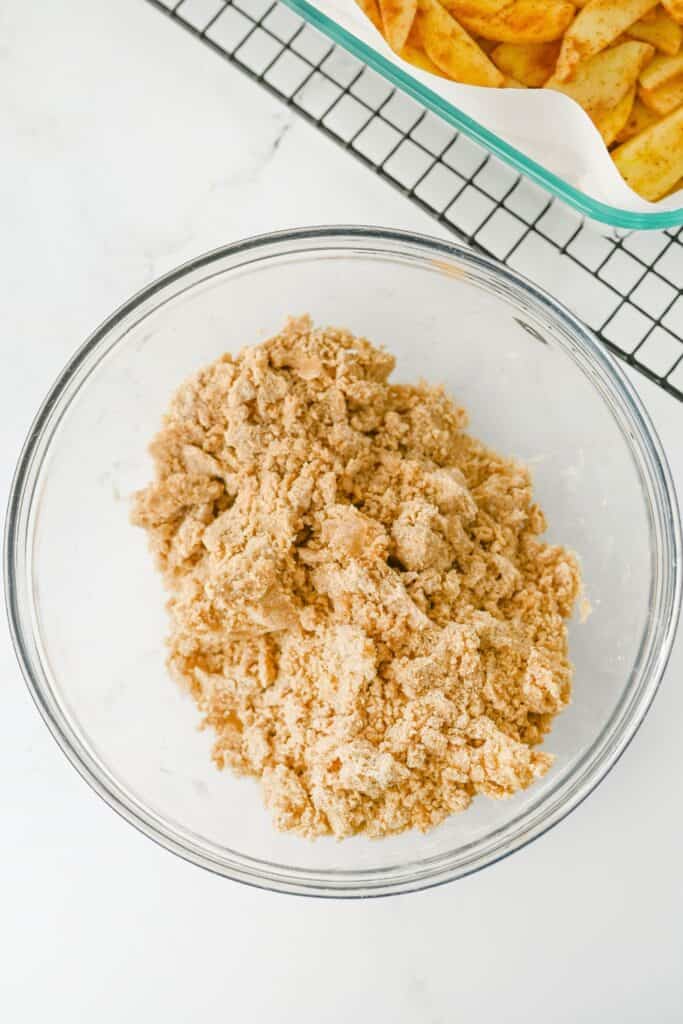 Overhead view of cinnamon apple crumble topping mixed into coarse buttery crumbs in a glass bowl. A pan of spiced apple slices sits nearby ready to be covered with the crumble topping before baking.