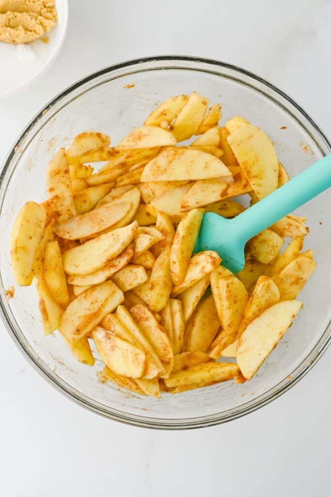 Sliced apples coated with cinnamon and spices in a glass mixing bowl for apple pie bars filling. A turquoise spatula rests among the seasoned apple slices ready to be layered over the crust.