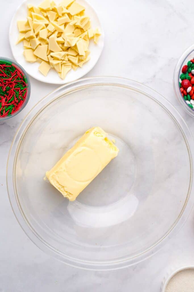 Stick of softened butter in a clear glass mixing bowl, surrounded by ingredients for Christmas cookies including chopped white chocolate, red and green sprinkles, and holiday M&Ms.