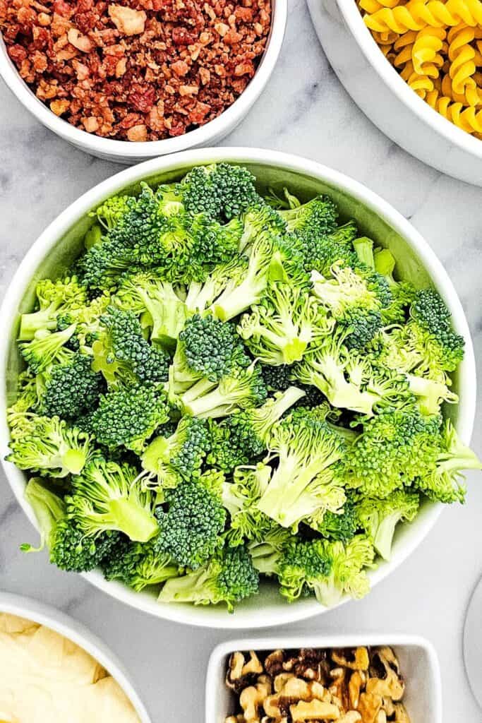 Bowl of fresh, chopped broccoli florets on a marble countertop, surrounded by other ingredients like bacon bits, rotini pasta, mayonnaise, and walnuts, prepared for a cold pasta salad.
