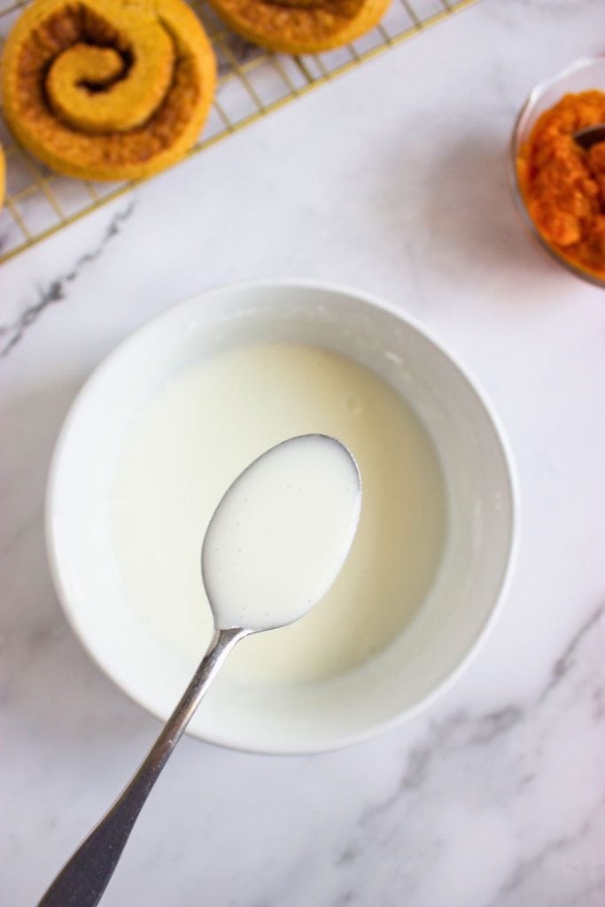 Spoon lifting a drizzle of smooth white glaze from a bowl, ready to be poured over cinnamon roll pumpkin cookies cooling on a rack in the background. A bowl of pumpkin purée sits nearby on the marble surface.