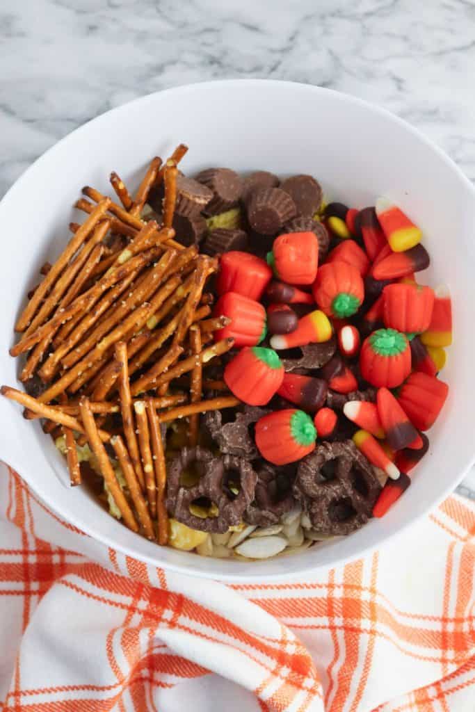Large white mixing bowl filled with Harvest Chex mix ingredients, including pretzel sticks, mini peanut butter cups, candy pumpkins, candy corn, chocolate-covered pretzels, and cereal pieces, resting on an orange and white plaid cloth on a marble surface.