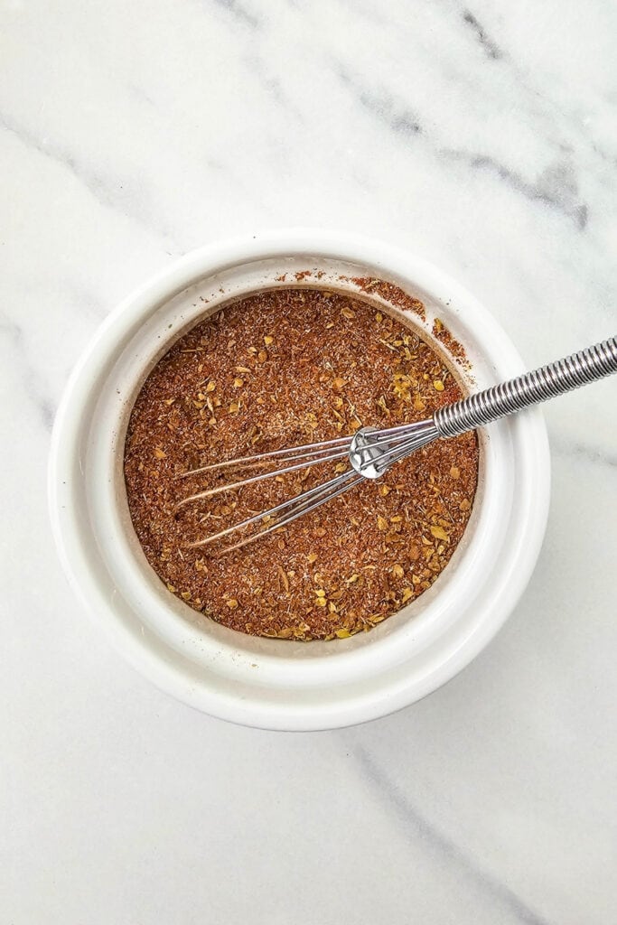 Homemade fajita spice mix fills a small white bowl on a marble countertop. A miniature metal whisk rests in the reddish brown blend of ground spices and dried herbs.