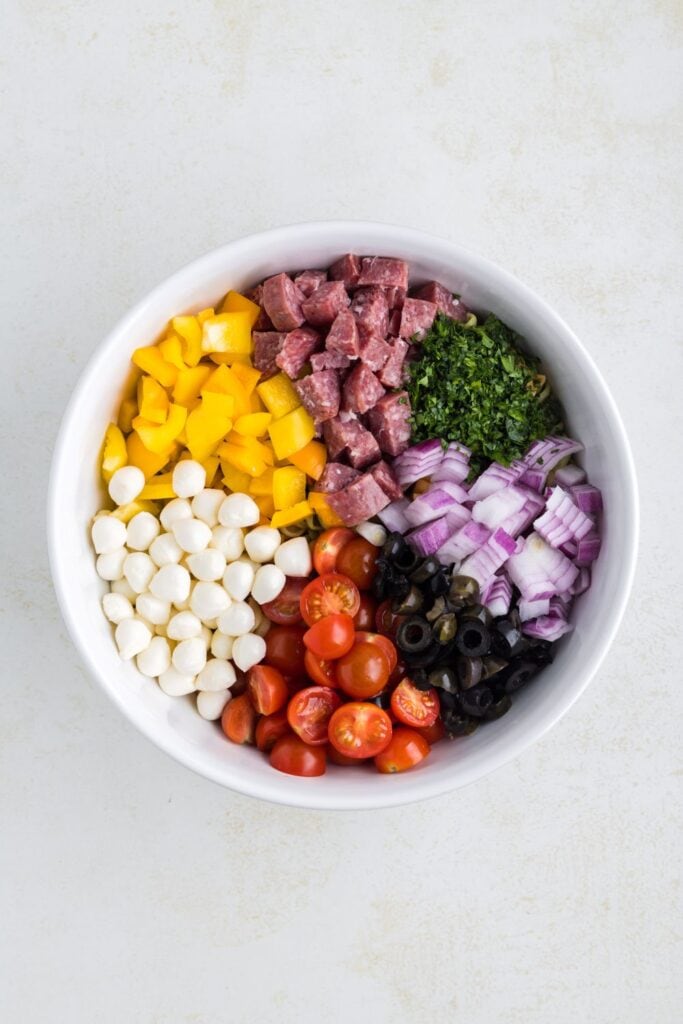A large white ceramic bowl filled with sections of cut up ingredients for zesty Italian pasta salad including cherry tomatoes, yellow peppers, parsley, salami, red onion, black olives, mini mozzarella balls.