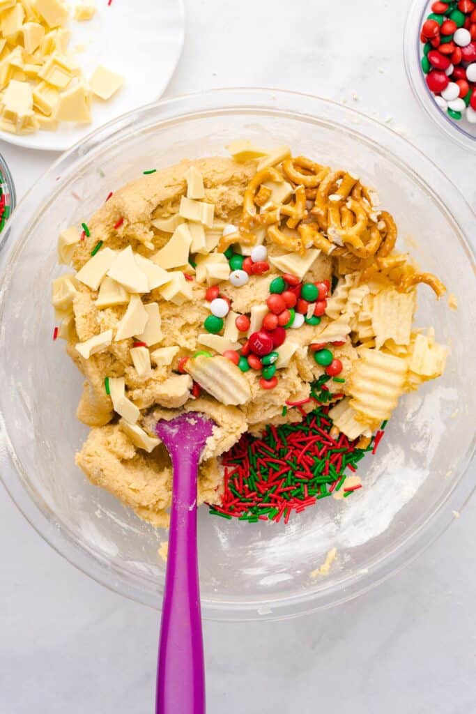 Bowl of cookie dough with holiday mix-ins on top, including red and green sprinkles, white chocolate chunks, Christmas M&Ms, pretzel twists, and potato chips, with a purple spatula ready to stir.